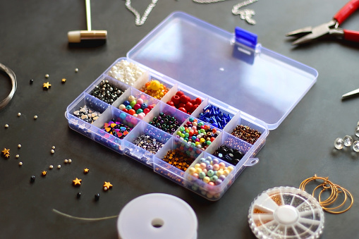 Box with colorful beads, string, wire, chain, scissors, pliers and hammer on dark background. Various jewelry making supplies. Selective focus.