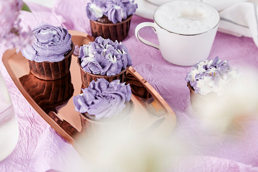 Aesthetics festive sweet table. Purple french cupcakes with cup of coffee among lilac flowers. Feminine lifestyle.