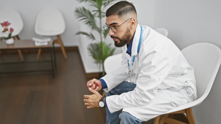 Handsome bearded man in lab coat sitting contemplatively in a modern clinic waiting room, exhaling a deep breath of relief.