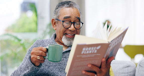 Elderly man, book and coffee on sofa with smile, reading or relax in retirement in home living room. Senior person, literature and happy with tea cup for knowledge, thinking and drink on lounge couch