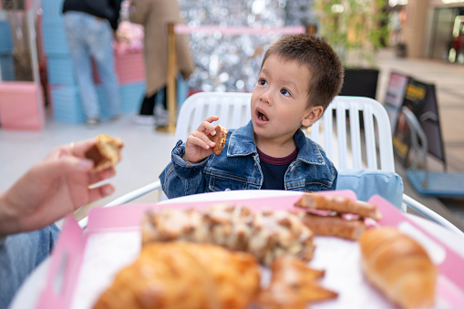 Charming two and a half year old boy, dressed in a stylish denim jacket, enjoys a delightful breakfast at an outdoor bakery table. His eyes light up with joy as he indulges in a selection of delicious pastries and croissants