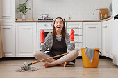 Overjoyed happy cheerful woman wearing apron and rubber gloves housewife cleaning floor with steam mop in kitchen at home clenched fist rejoicing to finish her household chores
