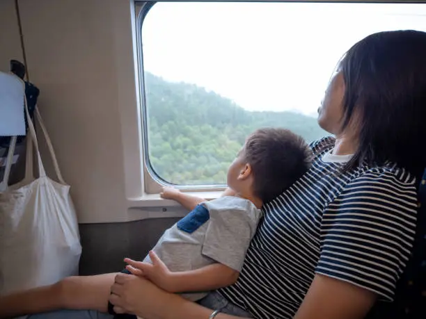 In a heartwarming scene, toddler sitting contentedly on his mother's lap as they embark on a train adventure, gazing out the window at passing landscapes. Perfect for projects celebrating family travel and the simple joys of shared experiences