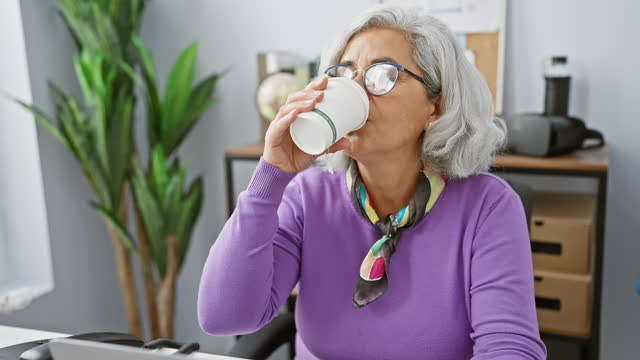 Mature woman in office enjoying coffee break, exuding casual professionalism and comfort, surrounded by plants and work-related objects.