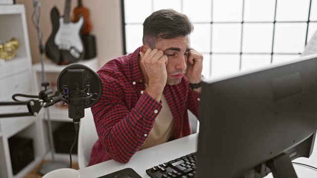 Stressed out, portrait of a young hispanic man, an upset musician facing problem with online performance while sitting in his music studio, somber interior reflecting his mood