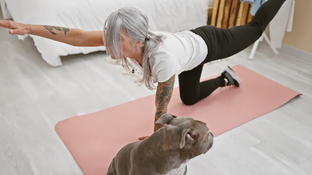 Sporty middle-aged woman, grey-haired and fit, trains yoga with her beloved dog, stretching her back in the relaxing ambiance of her bedroom