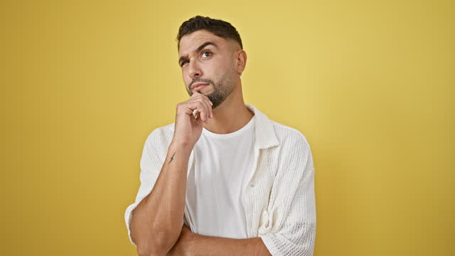 Young arab man, his finger on chin, standing, wondering with concentrated thought, puzzled, looking up in doubt against a yellow isolated background