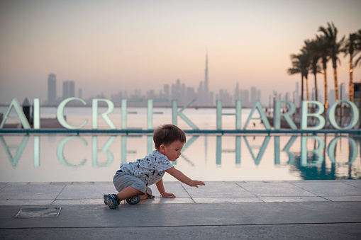 Witness the charm of a little explorer as a baby boy crawls in front of the Dubai Creek Harbor sign, determined to reach an empty bottle on the floor. The backdrop of the iconic Dubai Creek Harbor, with its city skyline, palm trees, and reflections in the water, adds a touch of magic to this endearing moment