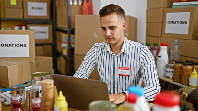 A handsome young adult man works attentively on a laptop at an indoor donation center surrounded by boxes and food supplies.
