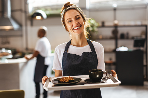 Happy waitress in a coffee shop holds a tray of delicious food, ready to serve customers and provide an excellent dining experience. Female server displaying a welcoming smile and a positive demeanour.