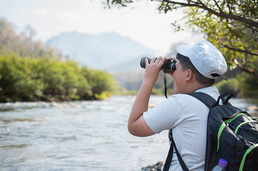 Asian boy wearing white t-shirt holding a binoculars sitting on stone by the river flowing down from the mountains in national park to observe fish in the river and birds on tree branches and on sky.