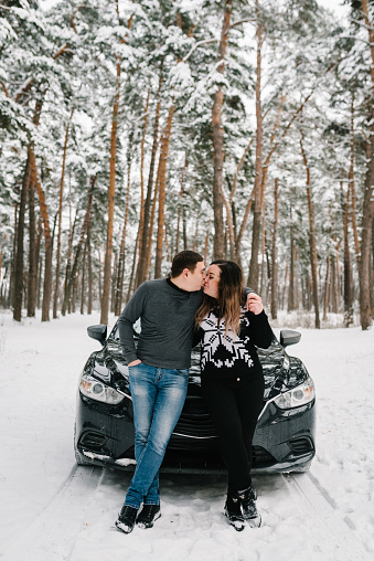 Couple kissing and leaning at parked modern car in snowy park looking at camera. Lovely man hug woman sitting on hood car of winter forest together. Nice couple has weekend at country at winter time.