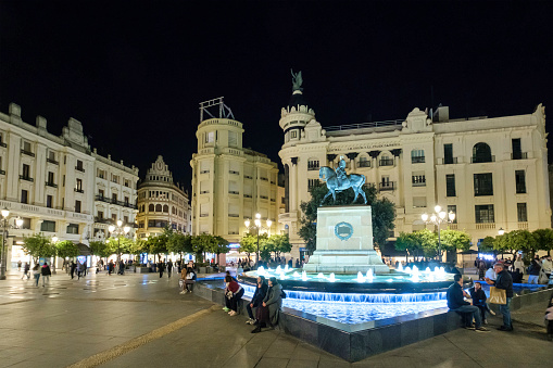 People sitting on the edge of the fountain where stands the equestrian monument to Gonzalo Fernández de Córdoba, work by Mateo Inurria in 1923. The location is the Plaza de las Tendillas, one of the popular squares in the Cordoba downtown.