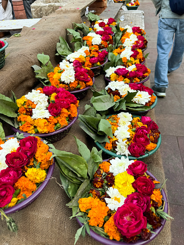 Stock photo showing close-up, elevated view of religious offerings of bowls of scented leaves and flower heads including, roses and marigolds, in rows on burlap covered tiered shelves outside a temple.