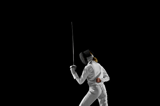 Female fencer stands at ready, her blade held high, poised to strike, but she waiting, her opponent's every move calculated against black studio background. Concept of professional sport, championship