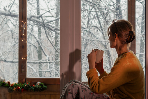 Young woman in peach homemade suit sits by window drinking coffee, enjoying winter landscape with snow-covered trees at Christmas time.