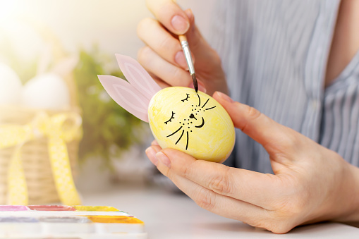 Happy Easter preparation. Young woman draws a cute face with a mustache on the eggshell of a yellow painted egg with pink bunny ears with a brush and paints. Close-up. Spring holiday.