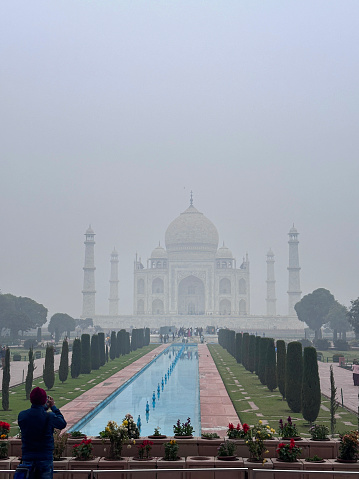 Stock photo showing Agra’s world-famous Mughal Taj Mahal, an imposing tomb mausoleum which dates back to the 17th century. The elaborate inlaid white marble architecture facade is flanked by four lighthouse-shaped towering minarets, being crowned by a series of large onion domes. Located in Agra, Uttar Pradesh, India, this Taj Mahal photo was taken early in the morning obscured by polluted foggy mist, air pollution caused by car exhaust fumes and factory emissions, with crowds of tourists taking selfies and photographs on their mobile phones.