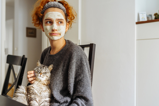 gray fluffy tabby cat sits in arms teenage girl, with red hair tied with hairband, wearing cosmetic clay mask