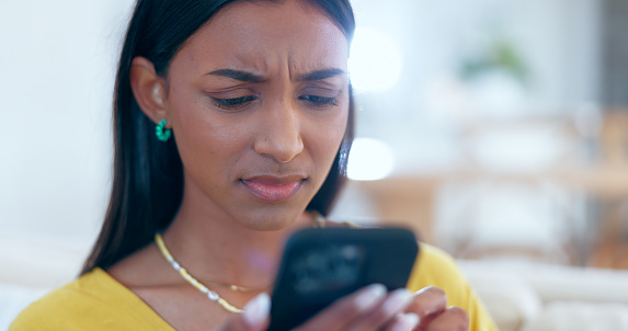 Confused, doubt and woman networking on a phone on sofa in the living room reading bad news. Upset, mad and young Indian female person scroll on social media or the internet with cellphone at home.