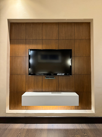 Stock photo showing close-up view of wall mounted flat screen television on bracket with hidden cables behind wood panel wall. The television screen is above a floating shelf with under shelf lighting.