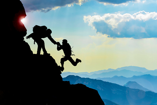 There are two climbers at the top of the mountains. They reach out and help each other on the cliff. They make an effort to reach the summit. They motivate each other by supporting each other.