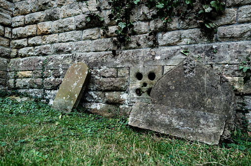 Old gravestones seen leaning against a stone wall of an old, medieval english church. The stones have been damaged and need to be repaired by a stone mason.