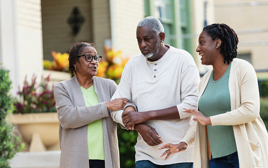 A senior African-American couple taking a walk with their adult daughter. Mother and daughter are conversing with each other, smiling. The father is in the middle, looking ahead with a serious expression. The seniors are in their 70s.