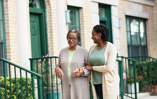 A senior African-American woman walking arm in arm, holding hands with her adult daughter down a sidewalk in a residential neighborhood in the city. Mother is in her 70s and her daughter is a mature woman in her 40s.