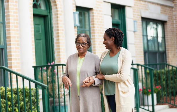 Senior woman walking arm in arm with daughter in city A senior African-American woman walking arm in arm, holding hands with her adult daughter down a sidewalk in a residential neighborhood in the city. Mother is in her 70s and her daughter is a mature woman in her 40s. locking-arms stock pictures, royalty-free photos & images