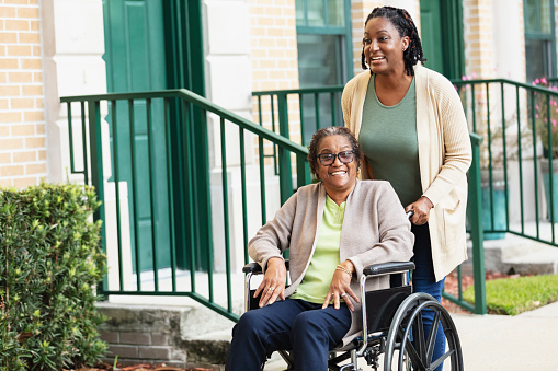 A caregiver pushing a senior African-American woman in a wheelchair on a city sidewalk in a residential neighborhood. The caregiver is the senior's adult daughter.
