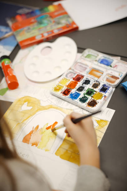 Two schoolgirls sitting at table, painting with water colors and having fun. Focus on the painting on the right