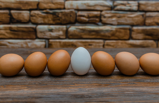 The photograph shows seven eggs arranged in a row on a wooden table in front of a stone wall. One of the eggs is white, the rest are brown. The white egg stands out among the brown ones and attracts attention. The lighting is soft and natural, emphasizing the textures of wood and stone.