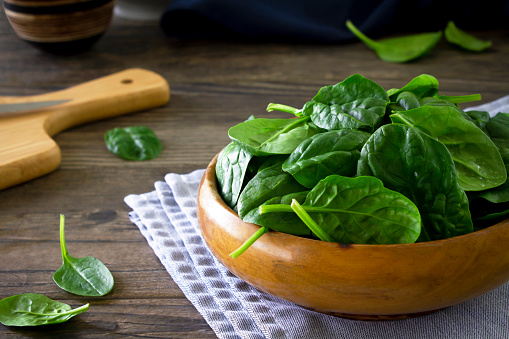 Close up shot of a bowl of fresh spinach leaves in a rustic bowl, on a wooden table with a chopping board.