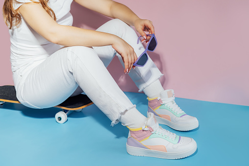 No face female in white outfit and retro style high-top multicolor sport sneakers shoes with sunglasses sitting on skateboard on blue pink background. Vintage retro fashion style of 80s - 90s vibes.