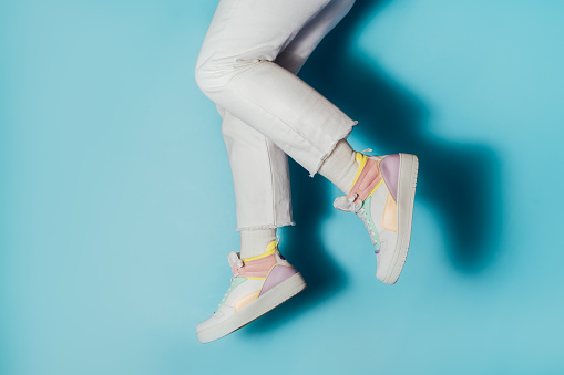 Close up female legs in white jeans and retro style high-top multicolor sport sneakers shoes jumping in the air on blue background. Pastel candy colors, vintage retro style of 80s - 90s vibes