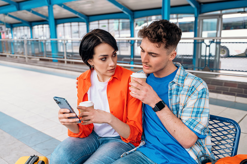 a young couple traveling together during their honeymoon, sitting in Interchange, waiting for bus, drinking coffee, chatting, reading news. Traveling together concept