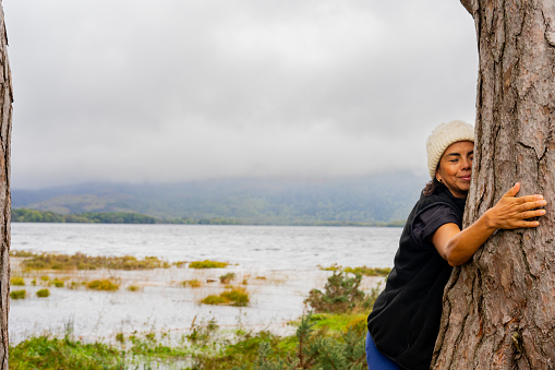 Latina woman with winter hat enjoying a moment of special connection with nature hugging a large pine trunk next to a lake in Ireland