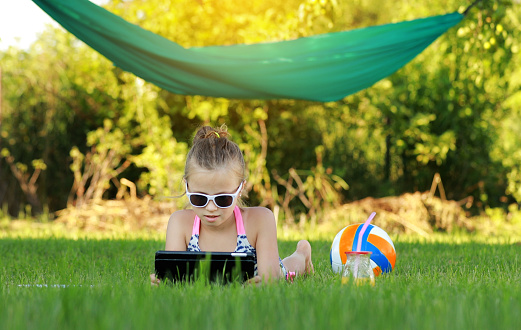 kid girl is eating piece of pizza and playing on laptop lying on grass on summer sunny day. the child has lunch after active games. hammock and ball on the background