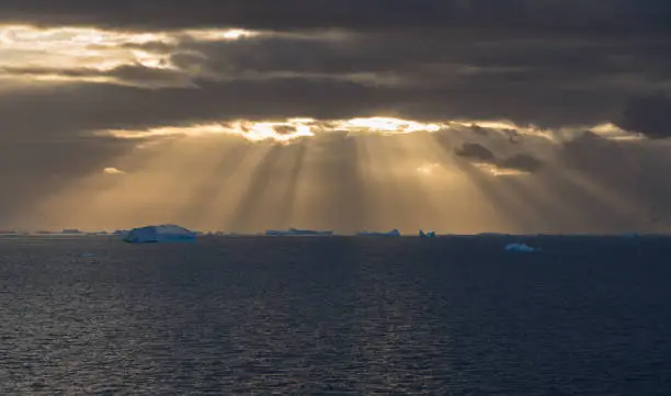 Icebergs in the distance illuminated by light rays breaking through the clouds.