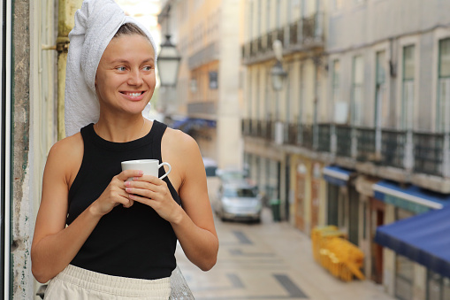 Young beautiful woman in a towel on head is drinking coffee or tea and smiling cute standing on balcony on ueropean street background. lifestyle morning. Concept of domestic or traveling lifestyle