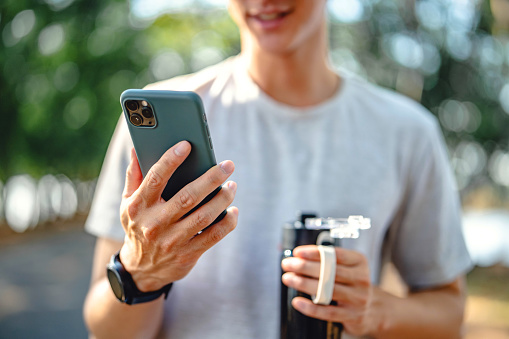 Close-up hand of man holding mobile phone and bottle of water at public park, Male using smartphone and wireless earbuds, checking mobile fitness application.