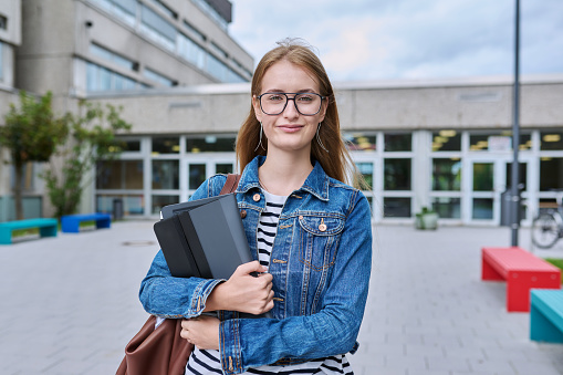 Portrait of teenage girl high school student, smiling female in glasses with backpack holding digital tablet, outdoor near educational building. Adolescence, education, knowledge concept