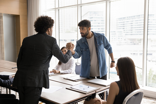 Two office competitors men fighting in arm wrestling battle over table, arguing at diverse team meeting, expressing disagreement, having problems with teamwork, competing for leadership