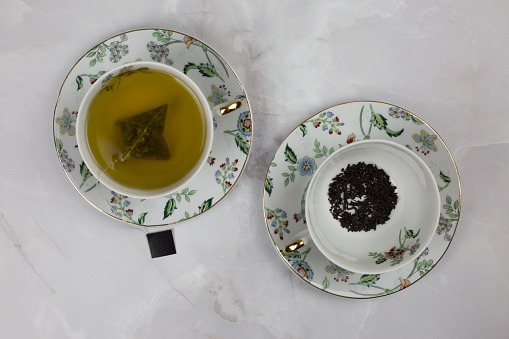 Cups and saucers with a tea bag of green tea and black tea leaves on a white table. Choice of hot drink, black or green tea
