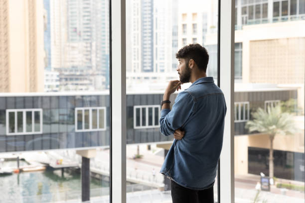 istock Thoughtful serious handsome young Arab man standing at large window 1917794810