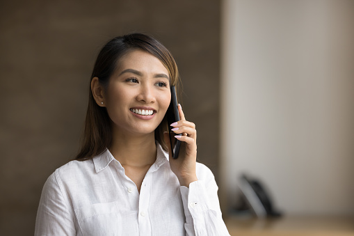 Cheerful young beautiful Asian manager talking to customer on mobile phone, smiling, enjoying connection, conversation, making job call, using wireless technology for work communication