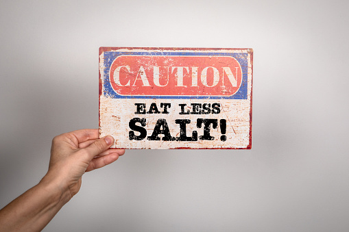 Eat Less Salt. Metal warning sign in a woman's hand on a white background.