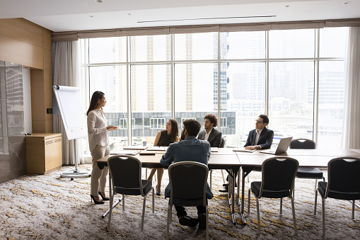 Young Asian business leader woman presenting project to diverse team of managers, telling management strategy to colleagues, talking to workgroup in modern office boardroom interior. Full length shot