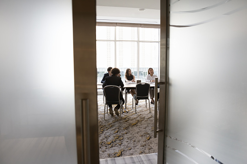 Diverse young business team sitting at large table in meeting room, talking, discussing collaboration on project, brainstorming on creative ideas. Full length candid shot, door view